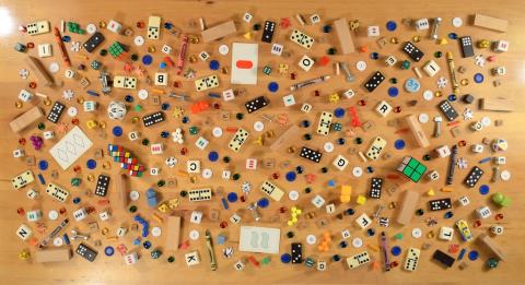 Many small items, such as dominoes, letter tiles from board games, dice, and puzzle pieces, are scattered on a wooden tabletop.