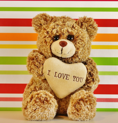 Little brown bear sitting in front of a red, white green and orange striped background holding an "I love you" heart