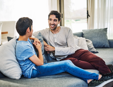 A photo of a father and his preteen son sitting on a couch and talking.
