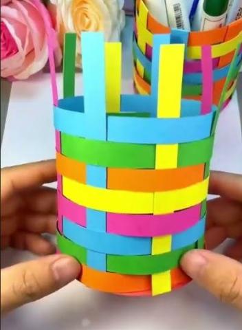 The hands of a white person hold an incomplete woven paper pencil holder. The pencil holder shows varying rows and layers of orange, green pink, blue, and yellow paper. In the background is a completed pencil holder with various office supplies inside it.