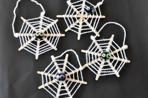 Four spiderwebs made of popsicle sticks and white yarn. In the middle of each is a pompom spider. The four webs sit on a black paper surface.