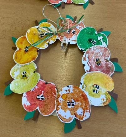 An autumn wreath. The wreath is ringed with apple stamps of various colors, all with stems and green leaves. There is a loop of brown twine at the top of the wreath.