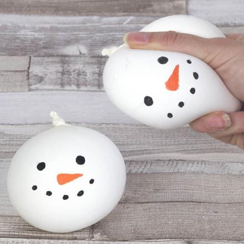 Two white balloons decorated to look like snowman heads. One of the balloons is being squished by a white person's hand to demonstrate that it is a stress ball.