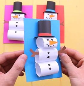 A white person's hands hold a piece of blue paper, on which are three pieces of white paper curled into tubes and decorated to look like a snowman. The snowman is wearing a black top hat, also made from rolled paper. In the background there are similar crafts on pink and red paper respectively.