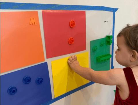 Orange, red, blue, yellow, and green construction paper are taped up behind transparent contact paper, sticky-side-out. A toddler is placing a small yellow object on the yellow paper. Other small objects are also on the contact paper, also sorted by color.
