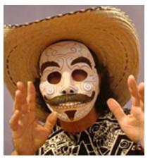 Performer Zarco Guerrero is pictured wearing a sombrero and highly decorated white mask with thick black eyebrows, mustache, and goatee. The mask is grinning toothily, and there are colorful swirls all over the mask.